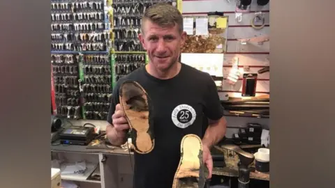 Andy Smith A man wearing a black t-shirt and carrying damaged shoes in a workshop with keys in the back.