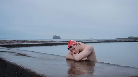 Jennifer Charlton Male swimmer in red swimming cap leaning on concrete beside outdoor pool, looking at camera, against gray background.