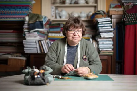 Jennifer Charlton A sad woman with glasses and a measuring tape sits at a table, surrounded by piles of books, and looks at the camera.