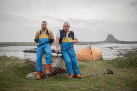 Jennifer Charlton Two fishermen in blue overalls lean on either side of the bow of a small fishing boat, anchored on the grass by the beach, with a small house far out on the water.