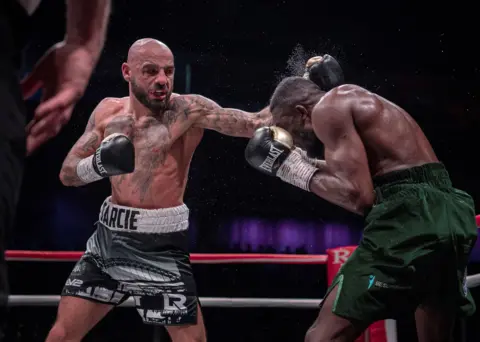 Jennifer Charlton Bald, a bearded boxer with chest and arm tattoos and a menacing face, lands a left punch to her opponent's head.