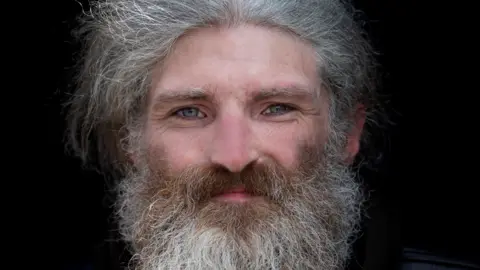 Jennifer Charlton A man with long gray hair and a beard and piercing blue eyes looking at the camera.