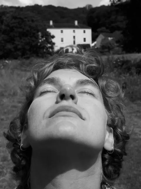 Mohamed Hassan Black and white photograph of a woman closing her eyes while standing in front of a large country house 