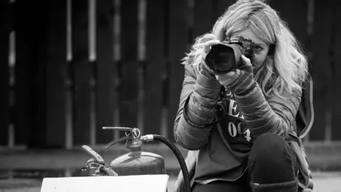 Jennifer Charlton Photographer Jennifer with long blonde hair in a warm jacket and gloves looks into the lens in this black and white photo.